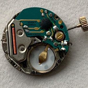 Puede incluir: Primer plano de un movimiento de reloj con una placa de circuito verde. La placa tiene el texto "SEVEN 7 JEWELS" y "UNADJUSTED". El mecanismo del reloj tiene una corona dorada y una caja de metal plateado.