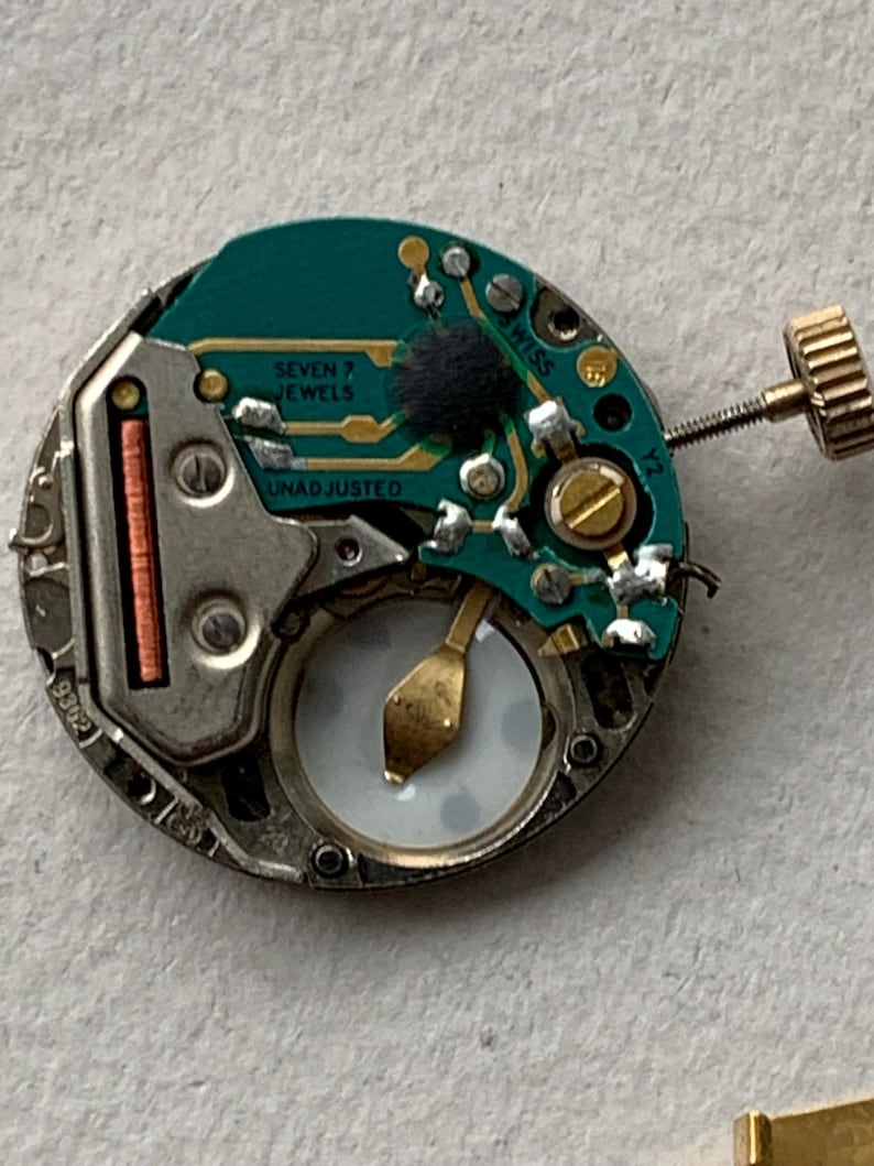 Puede incluir: Primer plano de un movimiento de reloj con una placa de circuito verde. La placa tiene el texto "SEVEN 7 JEWELS" y "UNADJUSTED". El mecanismo del reloj tiene una corona dorada y una caja de metal plateado.
