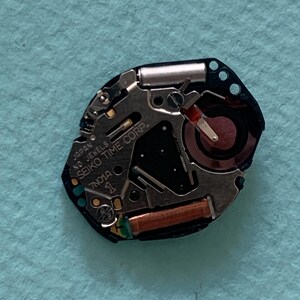 May include: A close-up of a watch movement with the text "JAPAN SEIKO TIME CORP." and "7N01A". The movement features silver, black, and copper components, including a small battery and a red-tipped hand. The background is a light blue surface.