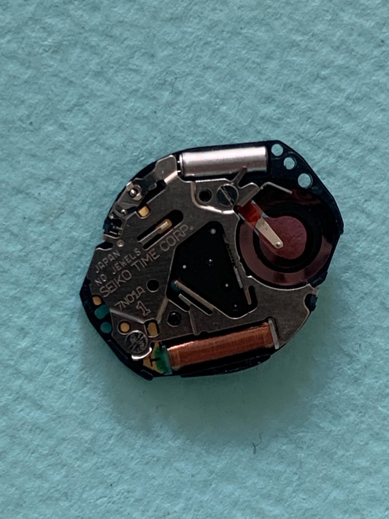 May include: A close-up of a watch movement with the text "JAPAN SEIKO TIME CORP." and "7N01A". The movement features silver, black, and copper components, including a small battery and a red-tipped hand. The background is a light blue surface.