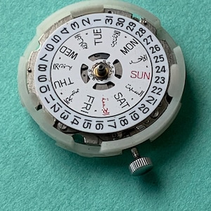 May include: A watch movement with a white dial featuring Arabic and English day and date indicators. The dial has a central mechanism and a small, silver-colored crown. The background is a solid teal color.