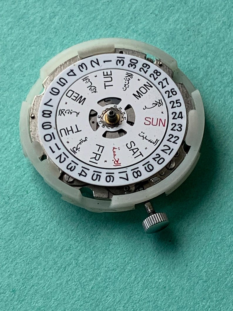 May include: A watch movement with a white dial featuring Arabic and English day and date indicators. The dial has a central mechanism and a small, silver-colored crown. The background is a solid teal color.