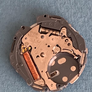 May include: Close-up of a watch movement with intricate mechanical components. The watch mechanism features a silver-colored base with a dark gray circular element and a copper-colored coil. Text on the movement reads "17 JEWELS JAPAN" and "S671L".