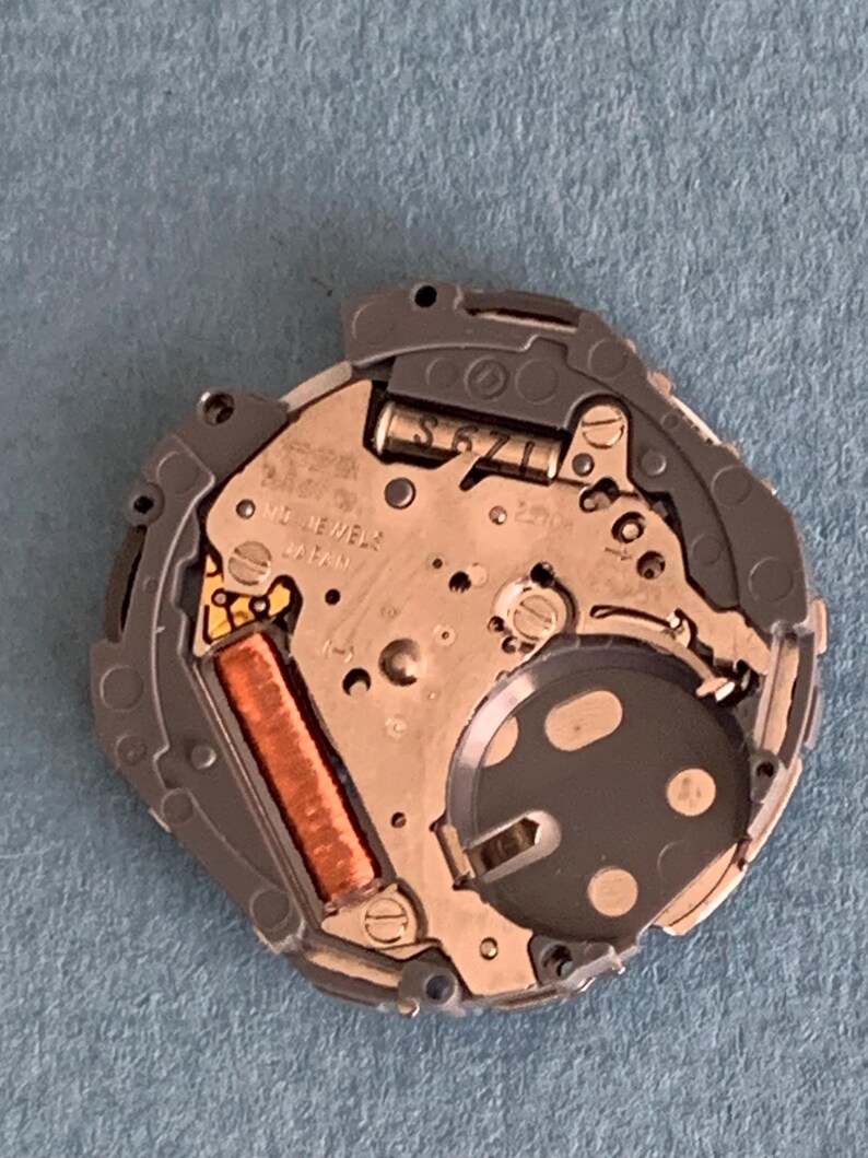 May include: Close-up of a watch movement with intricate mechanical components. The watch mechanism features a silver-colored base with a dark gray circular element and a copper-colored coil. Text on the movement reads "17 JEWELS JAPAN" and "S671L".