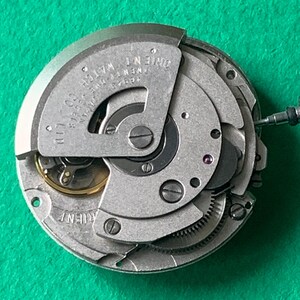 May include: Close-up of a mechanical watch movement. The silver-colored mechanism features gears, screws, and the text "ORIENT WATCH" and "TWENTY-ONE JEWELS". The watch movement is a complex assembly of small parts.