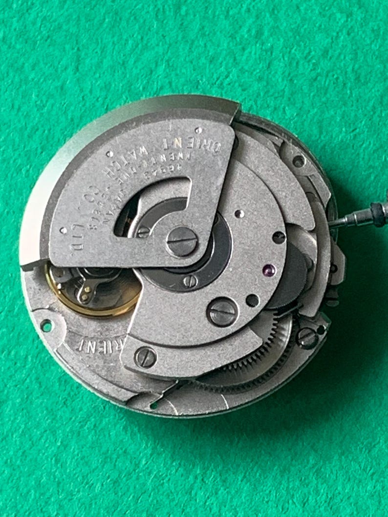 May include: Close-up of a mechanical watch movement. The silver-colored mechanism features gears, screws, and the text "ORIENT WATCH" and "TWENTY-ONE JEWELS". The watch movement is a complex assembly of small parts.