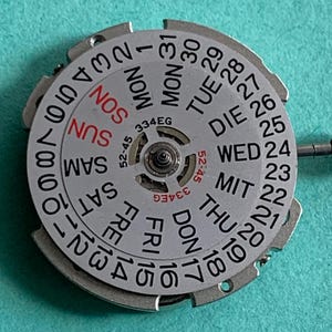 May include: A silver watch calendar wheel with black numbers and day abbreviations. The days of the week are in English, with "SUN" in red. The wheel has the text "334EG" and "52-45".
