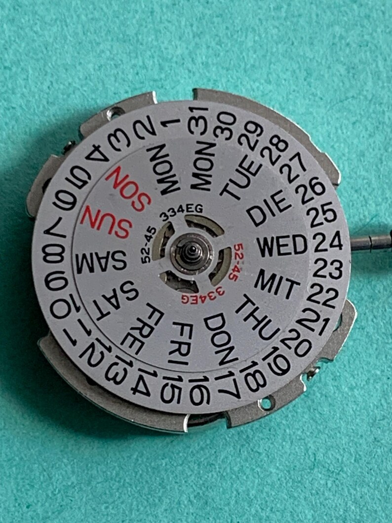 May include: A silver watch calendar wheel with black numbers and day abbreviations. The days of the week are in English, with "SUN" in red. The wheel has the text "334EG" and "52-45".