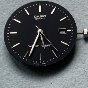 Puede incluir: Una esfera de reloj Casio Quartz negra con manecillas e indicadores plateados. La esfera del reloj tiene las palabras "CASIO QUARTZ" y "WATER RESIST" impresas. La fecha se muestra en una peque&ntilde;a ventana. El reloj est&aacute; marcado "JAPAN MOVT".
