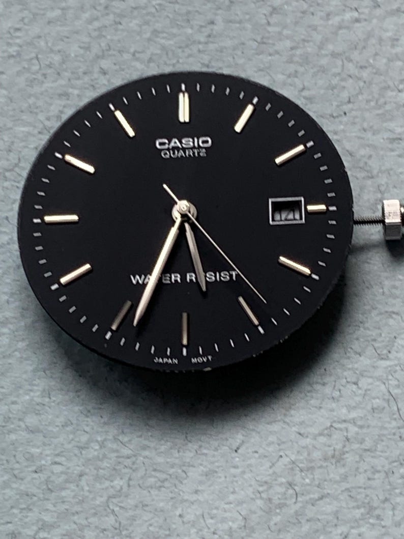 Puede incluir: Una esfera de reloj Casio Quartz negra con manecillas e indicadores plateados. La esfera del reloj tiene las palabras "CASIO QUARTZ" y "WATER RESIST" impresas. La fecha se muestra en una peque&ntilde;a ventana. El reloj est&aacute; marcado "JAPAN MOVT".