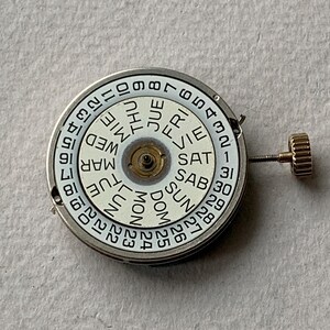 Puede incluir: Un movimiento de reloj con una esfera blanca que muestra los d&iacute;as de la semana y las fechas numeradas. La esfera tiene un marco de metal plateado y una corona dorada. Los d&iacute;as de la semana est&aacute;n en espa&ntilde;ol.