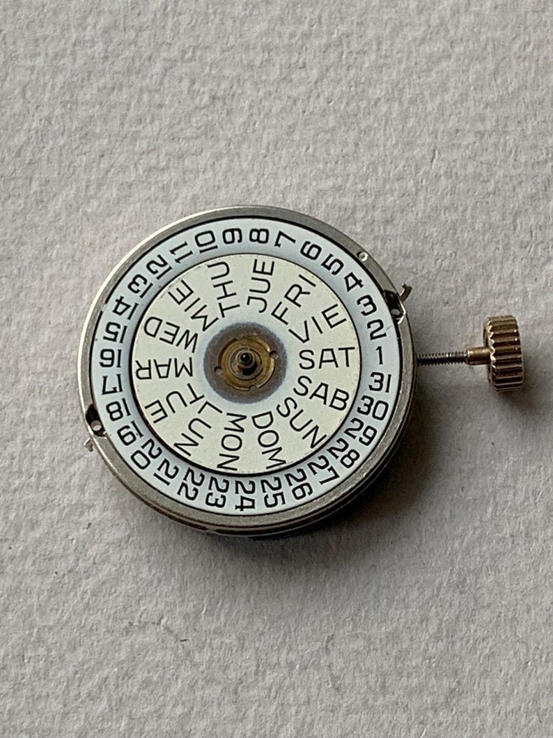 Puede incluir: Un movimiento de reloj con una esfera blanca que muestra los d&iacute;as de la semana y las fechas numeradas. La esfera tiene un marco de metal plateado y una corona dorada. Los d&iacute;as de la semana est&aacute;n en espa&ntilde;ol.