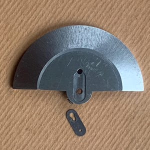 Pu&ograve; includere: Primo piano di due piccole parti metalliche argentate di orologio. Una &egrave; un pezzo semicircolare con un piccolo ingranaggio e un foro. L'altra &egrave; un piccolo pezzo oblungo con un foro. Entrambi sono su una superficie beige.