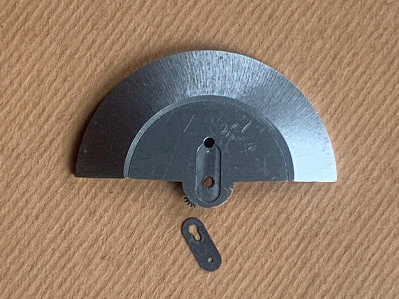 Pu&ograve; includere: Primo piano di due piccole parti metalliche argentate di orologio. Una &egrave; un pezzo semicircolare con un piccolo ingranaggio e un foro. L'altra &egrave; un piccolo pezzo oblungo con un foro. Entrambi sono su una superficie beige.