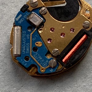May include: A detailed view of a watch movement, featuring a blue circuit board and gold-toned metal parts. The text "UNADJUSTED SWISS V8" and "ELEVEN JEWELS" are visible. Includes a small red coil and a black battery.