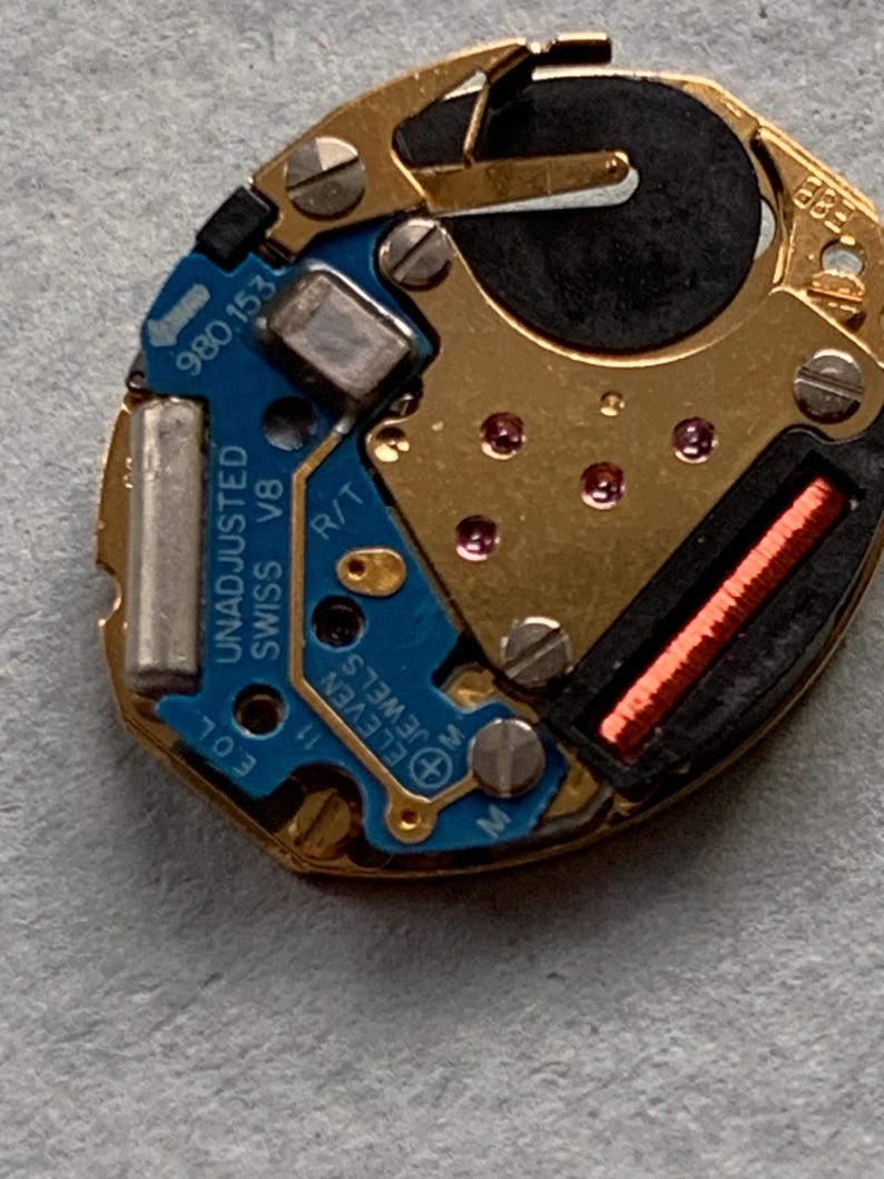 May include: A detailed view of a watch movement, featuring a blue circuit board and gold-toned metal parts. The text "UNADJUSTED SWISS V8" and "ELEVEN JEWELS" are visible. Includes a small red coil and a black battery.