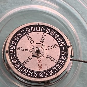 May include: Close-up of a watch movement with a silver-colored metal frame and a white dial. The dial features black numerals and the days of the week in German. The date is displayed around the outer edge.