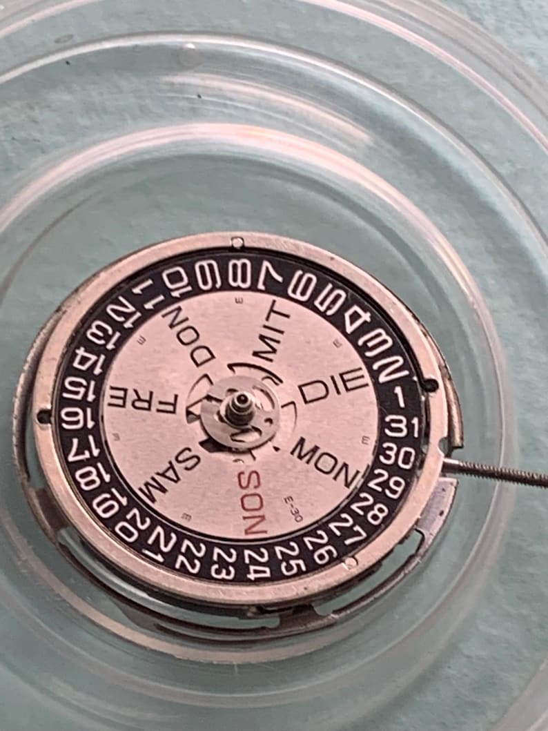 May include: Close-up of a watch movement with a silver-colored metal frame and a white dial. The dial features black numerals and the days of the week in German. The date is displayed around the outer edge.