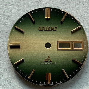 May include: A green and gold watch face with the text "ORIENT" and "21 JEWELS" printed on it. The watch face also has the text "JAPAN LM 4294-2367-06" printed on it.