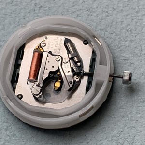 Puede incluir: Primer plano de un movimiento de reloj con una carcasa de pl&aacute;stico blanco. Los componentes internos incluyen una bobina de cobre, piezas met&aacute;licas plateadas y un tornillo peque&ntilde;o. El movimiento est&aacute; etiquetado como "MIYOTA CO. JAPAN 2115 UNADJUSTED NO JEWELS."