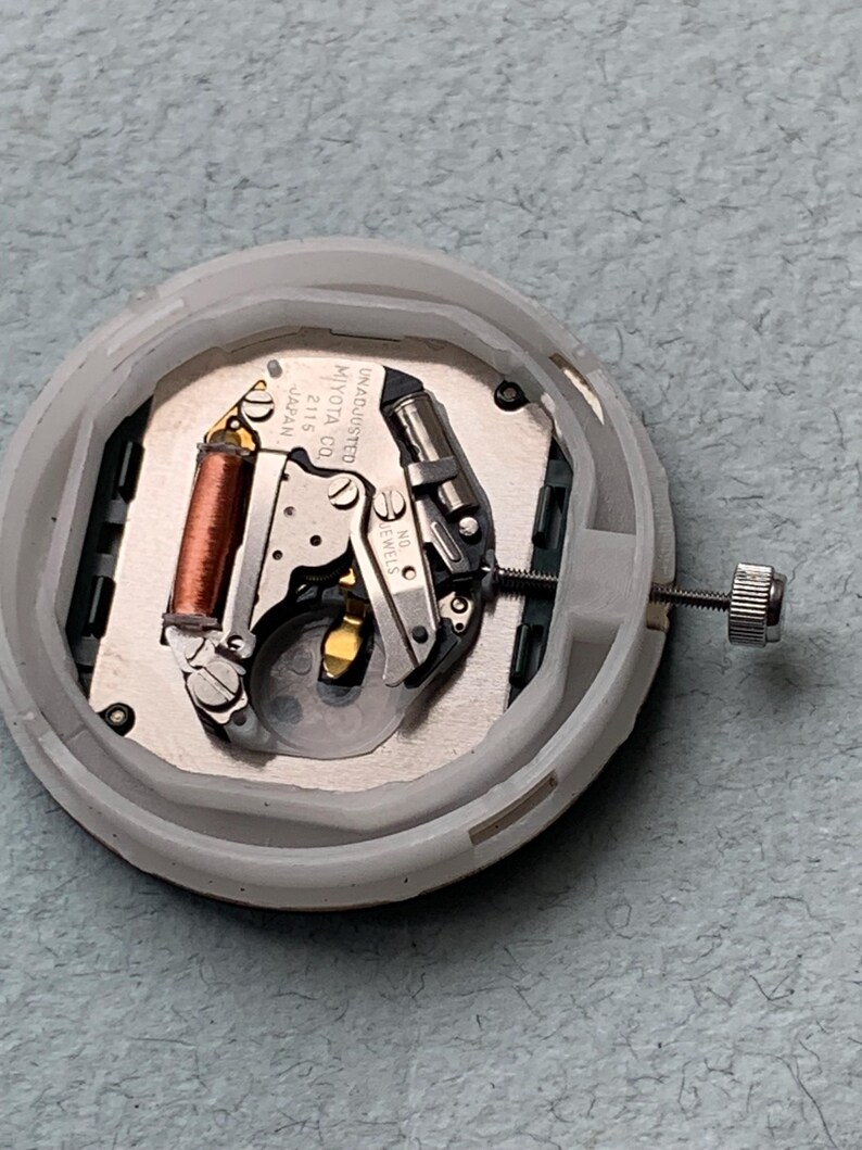 Puede incluir: Primer plano de un movimiento de reloj con una carcasa de pl&aacute;stico blanco. Los componentes internos incluyen una bobina de cobre, piezas met&aacute;licas plateadas y un tornillo peque&ntilde;o. El movimiento est&aacute; etiquetado como "MIYOTA CO. JAPAN 2115 UNADJUSTED NO JEWELS."