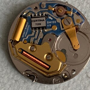 Może przedstawiać: Zbliżenie mechanizmu zegarka z misternymi detalami. Mechanizm zegarka zawiera niebieską płytkę obwodu, złote elementy i napis "CERTINA 729 SWISS V8". Mechanizm zegarka to złożony zesp&oacute;ł małych części.