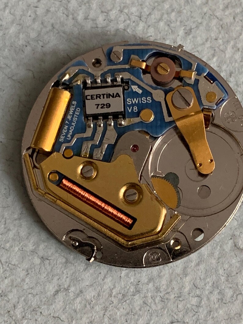 Może przedstawiać: Zbliżenie mechanizmu zegarka z misternymi detalami. Mechanizm zegarka zawiera niebieską płytkę obwodu, złote elementy i napis "CERTINA 729 SWISS V8". Mechanizm zegarka to złożony zesp&oacute;ł małych części.