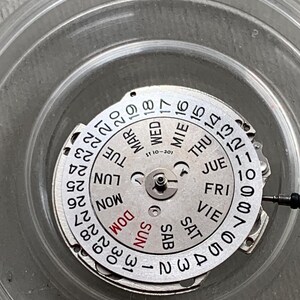 Pu&ograve; includere: Un movimento di orologio argentato con un quadrante circolare che mostra i giorni della settimana e la data. Il quadrante ha numeri e testo neri, con "SUN" in rosso. Il movimento &egrave; all'interno di un contenitore di plastica trasparente.