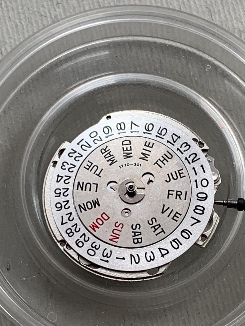 Pu&ograve; includere: Un movimento di orologio argentato con un quadrante circolare che mostra i giorni della settimana e la data. Il quadrante ha numeri e testo neri, con "SUN" in rosso. Il movimento &egrave; all'interno di un contenitore di plastica trasparente.
