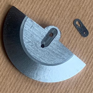 Pu&ograve; includere: Primo piano di componenti di orologi. Un pezzo di metallo semicircolare color argento con un bordo nero e una punta bianca, e un piccolo pezzo di metallo scuro con due fori. I pezzi sono su una superficie marrone chiaro.