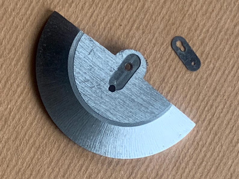 Pu&ograve; includere: Primo piano di componenti di orologi. Un pezzo di metallo semicircolare color argento con un bordo nero e una punta bianca, e un piccolo pezzo di metallo scuro con due fori. I pezzi sono su una superficie marrone chiaro.
