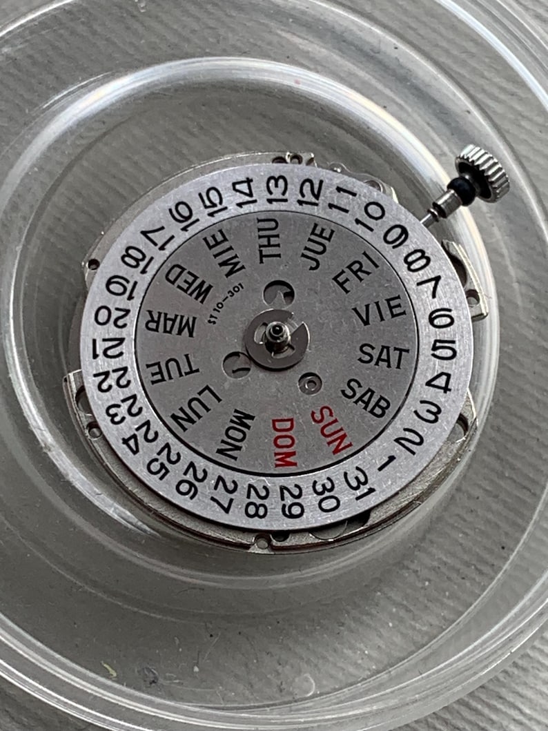 Pu&ograve; includere: Un movimento di orologio argentato con visualizzazione del giorno e della data. Il quadrante presenta numeri neri e abbreviazioni dei giorni in pi&ugrave; lingue. Una piccola corona &egrave; visibile sul lato. Il movimento &egrave; in un contenitore di plastica trasparente.