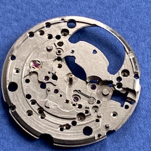 Puede incluir: Primer plano de un movimiento de reloj plateado con intrincados engranajes, agujeros y peque&ntilde;as joyas. Los componentes mec&aacute;nicos est&aacute;n dispuestos sobre una placa circular, mostrando la precisi&oacute;n del funcionamiento interno del reloj. El fondo es azul liso.