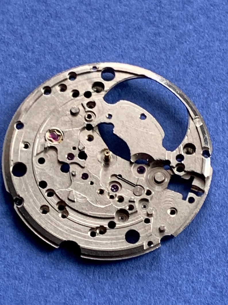 Puede incluir: Primer plano de un movimiento de reloj plateado con intrincados engranajes, agujeros y peque&ntilde;as joyas. Los componentes mec&aacute;nicos est&aacute;n dispuestos sobre una placa circular, mostrando la precisi&oacute;n del funcionamiento interno del reloj. El fondo es azul liso.