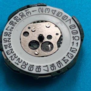May include: A close-up of a watch movement, featuring a white date dial with black numerals. The dial is set within a dark green frame. The internal mechanism includes silver gears and screws, and a central post.