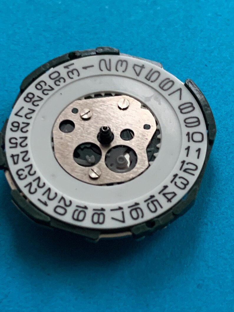 May include: A close-up of a watch movement, featuring a white date dial with black numerals. The dial is set within a dark green frame. The internal mechanism includes silver gears and screws, and a central post.