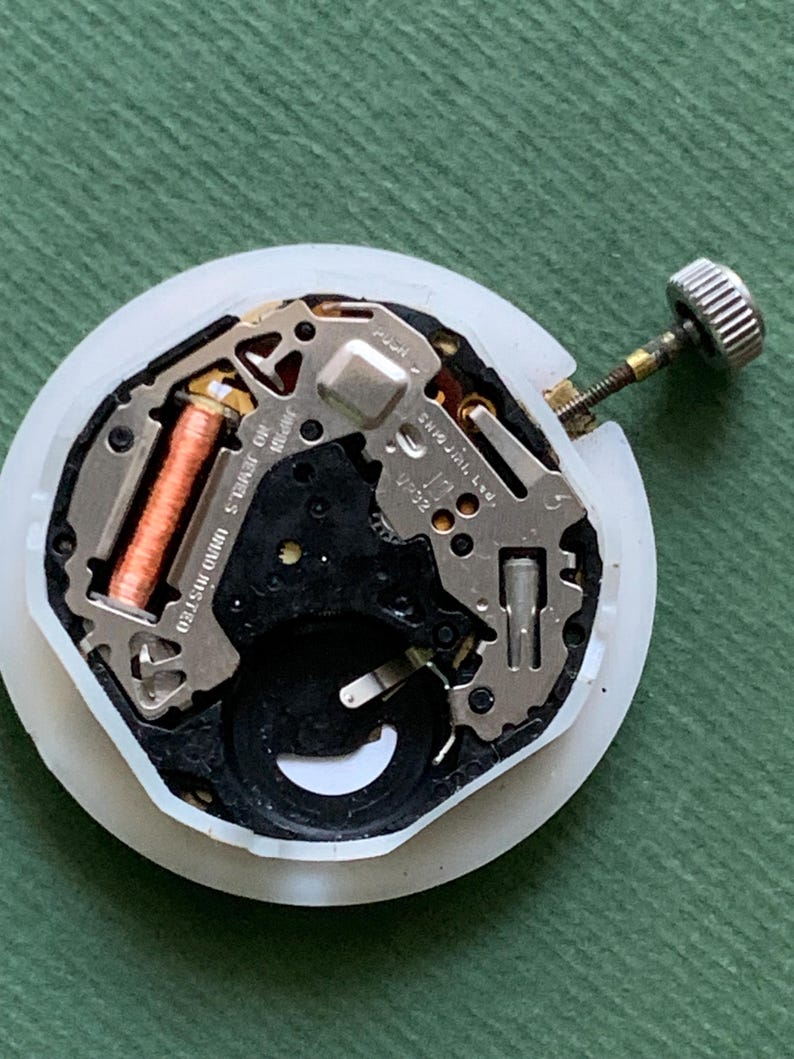 K&ouml;nnte beinhalten: Ein zerlegtes Uhrwerk mit einem wei&szlig;en Kunststoffgeh&auml;use. Die inneren Komponenten umfassen Metallplatten, eine Kupferspule und eine silberfarbene Krone. Das Uhrwerk ist mit "Japan" und "10 Jewels" beschriftet.