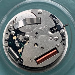 Op de afbeelding: Close-up van een uurwerk met diverse componenten, waaronder een batterij, tandwielen en een spoel. Het horlogemechanisme is zilverkleurig met de tekst "Vamee" en "Swiss AV" zichtbaar. Het uurwerk staat tegen een lichtblauwe achtergrond.