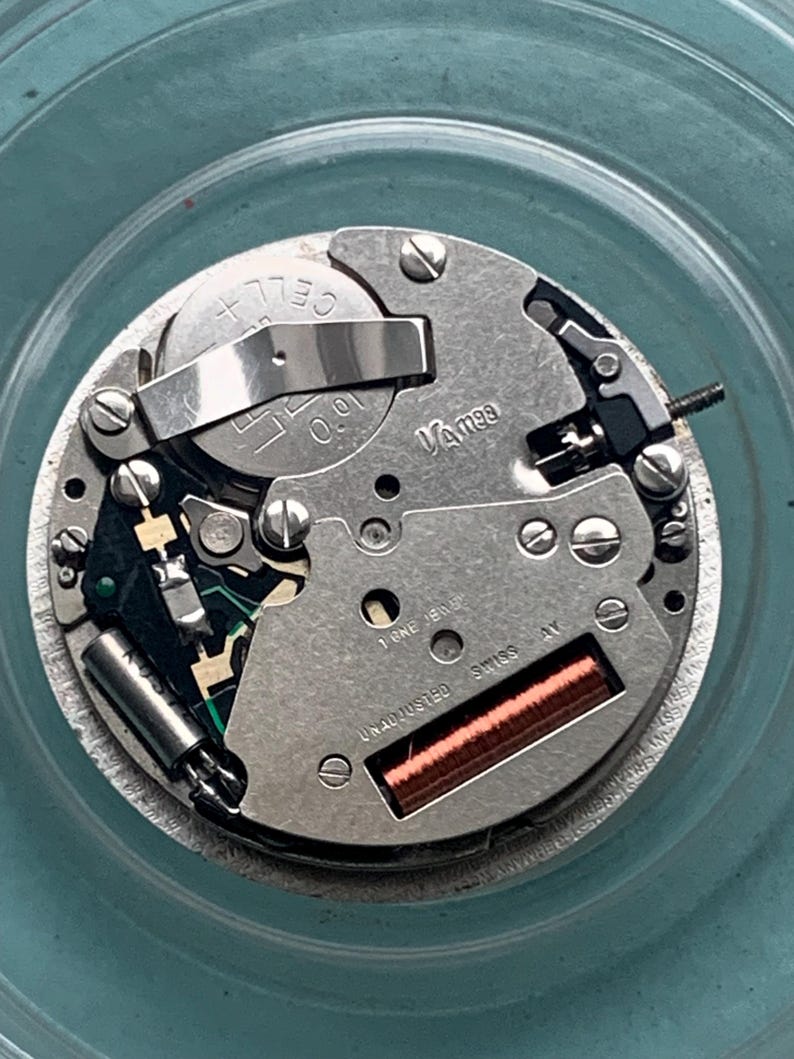 Op de afbeelding: Close-up van een uurwerk met diverse componenten, waaronder een batterij, tandwielen en een spoel. Het horlogemechanisme is zilverkleurig met de tekst "Vamee" en "Swiss AV" zichtbaar. Het uurwerk staat tegen een lichtblauwe achtergrond.