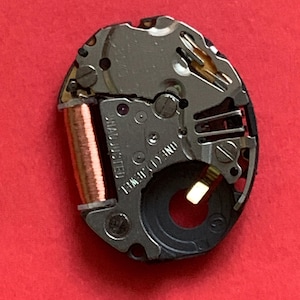 May include: Close-up of a watch movement on a red background. The movement is oval-shaped, with silver and copper components. The text "UNADJUSTED" and "ONE (1) JEWEL" are visible. The watch mechanism is a detailed example of watchmaking.