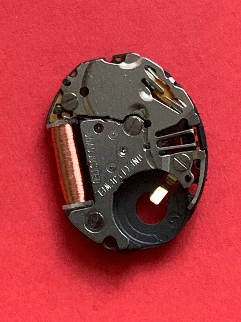 May include: Close-up of a watch movement on a red background. The movement is oval-shaped, with silver and copper components. The text "UNADJUSTED" and "ONE (1) JEWEL" are visible. The watch mechanism is a detailed example of watchmaking.