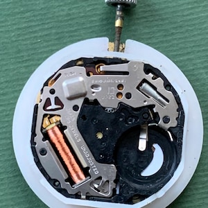 K&ouml;nnte beinhalten: Nahaufnahme eines Uhrwerks mit einem wei&szlig;en Kunststoffring. Zu den internen Komponenten geh&ouml;ren eine Kupferspule, eine schwarze Batterie und Metallzahnr&auml;der. Der Text "SHIOJIRI Ltd." und "JAPAN 0 JEWELS UNADJUSTED" sind sichtbar.