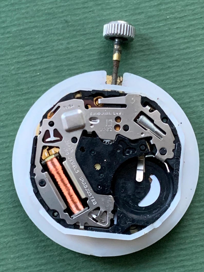 K&ouml;nnte beinhalten: Nahaufnahme eines Uhrwerks mit einem wei&szlig;en Kunststoffring. Zu den internen Komponenten geh&ouml;ren eine Kupferspule, eine schwarze Batterie und Metallzahnr&auml;der. Der Text "SHIOJIRI Ltd." und "JAPAN 0 JEWELS UNADJUSTED" sind sichtbar.