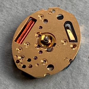 May include: A detailed view of a gold-coloured watch movement, showcasing intricate gears, coils, and small screws. The oval mechanism features a complex internal structure with various components and openings. The watch part rests on a grey surface.