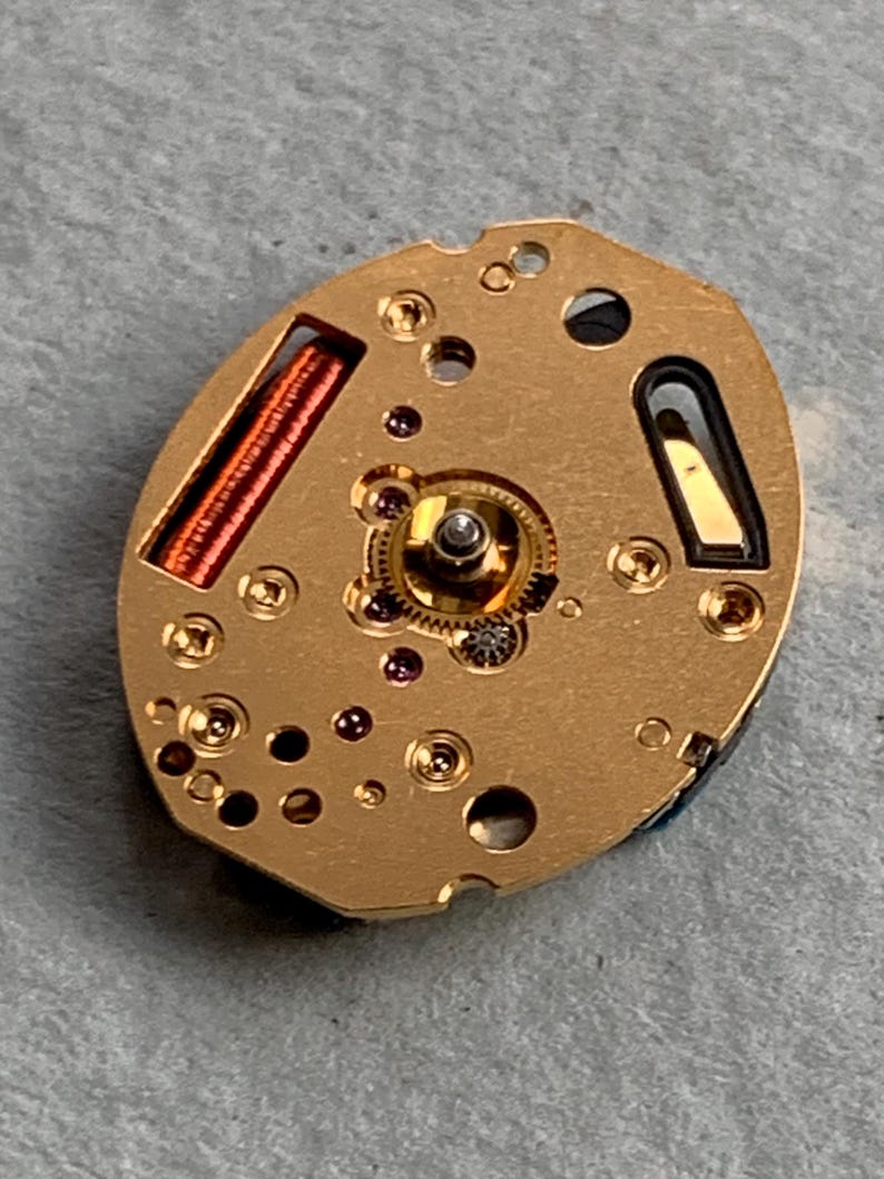 May include: A detailed view of a gold-coloured watch movement, showcasing intricate gears, coils, and small screws. The oval mechanism features a complex internal structure with various components and openings. The watch part rests on a grey surface.