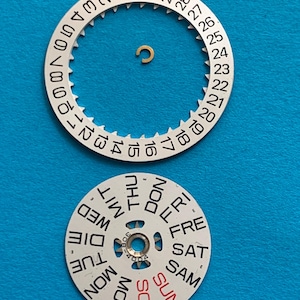 May include: Watch parts, including a silver-colored date wheel with numbers 1-31 and a day wheel with days of the week in black and red. A small gold-colored component is also present. The parts are on a blue background.