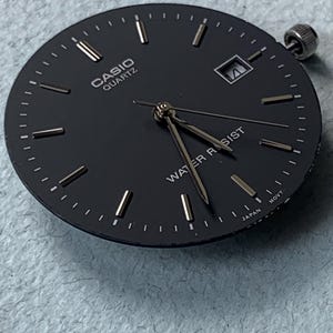 Puede incluir: Una esfera de reloj Casio Quartz negra con marcadores de hora y agujas plateadas. La esfera del reloj tiene las palabras "CASIO QUARTZ" y "WATER RESIST" impresas. El reloj tambi&eacute;n tiene una ventana de fecha y una corona plateada.