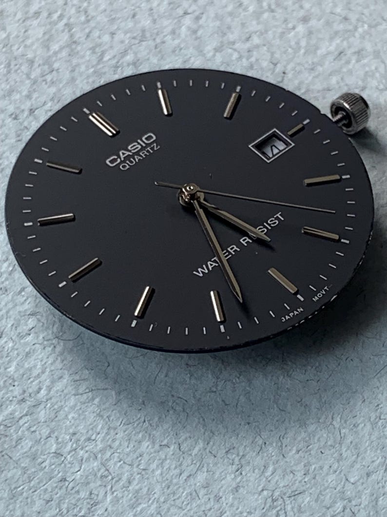 Puede incluir: Una esfera de reloj Casio Quartz negra con marcadores de hora y agujas plateadas. La esfera del reloj tiene las palabras "CASIO QUARTZ" y "WATER RESIST" impresas. El reloj tambi&eacute;n tiene una ventana de fecha y una corona plateada.