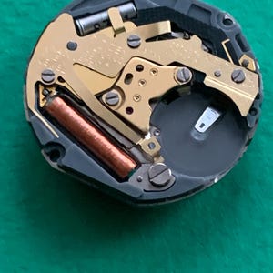May include: Close-up of a watch movement with gold-colored gears, a brown coil, and a black battery compartment. The intricate mechanism is set within a black frame, showcasing the internal components of a timepiece.