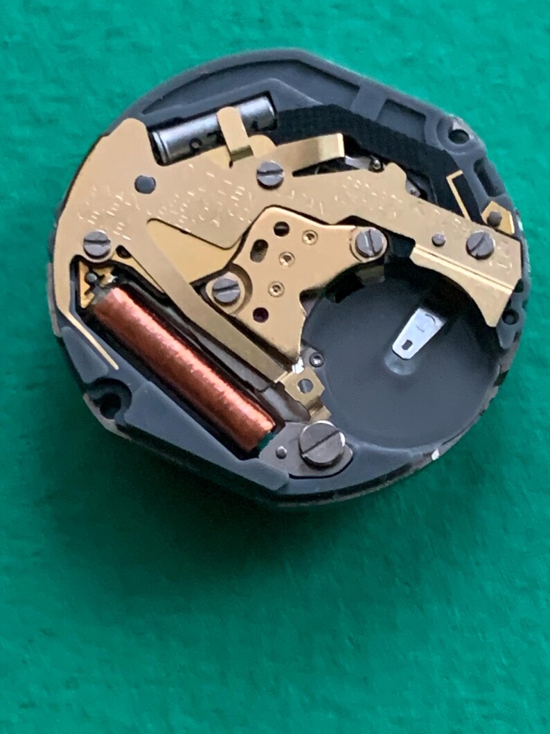 May include: Close-up of a watch movement with gold-colored gears, a brown coil, and a black battery compartment. The intricate mechanism is set within a black frame, showcasing the internal components of a timepiece.
